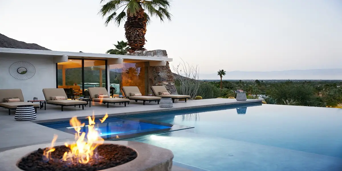 A modern outdoor patio at one of the best hotels in Palm Springs features a pool with an infinity edge, surrounded by lounge chairs and a fire pit in the foreground. A glass-walled building with warm interior lighting is to the left. Palm trees and a scenic view of mountains and valley are in the background.