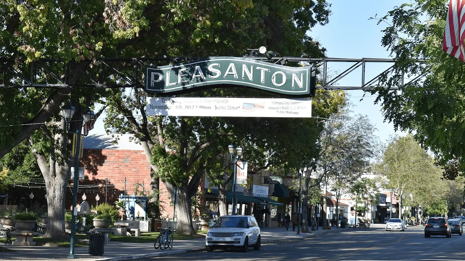 A street in Pleasanton, California, features a large overhead sign that reads "PLEASANTON" with trees and buildings lining the sidewalk. A few cars are parked along the street, and a banner advertising an event is attached below the sign. An American flag waves on the right, inviting you to visit East Bay's Tri-Valley.