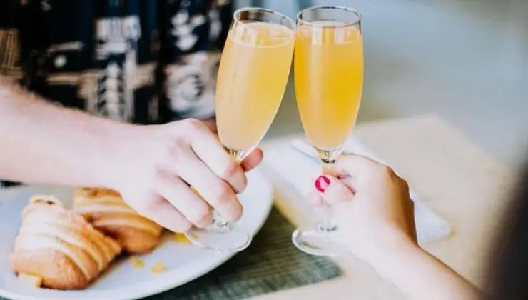 Two hands clink champagne flutes filled with a light orange drink, possibly mimosas, against a blurred background. In the foreground, pastries on a white plate are partially visible. The setting appears casual and festive—an ideal scene for enjoying the best breakfast Kauai has to offer.