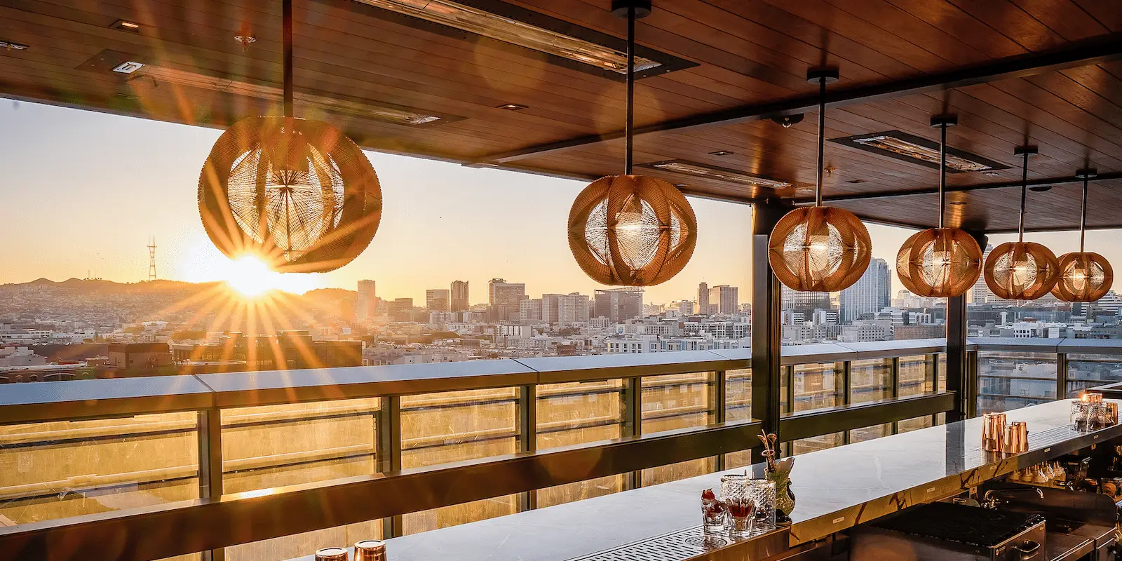 A modern rooftop bar at sunset with a city skyline view, perfect for the coolest rooftop dining experience in the Bay Area. The bar features a sleek counter with drinks and elegant, spherical pendant lights hanging from a wooden ceiling. The sun sets over distant buildings, casting a warm glow across the scene.