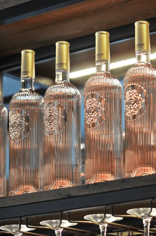 Several elegant, ribbed glass bottles of rosé wine from Provence with gold caps are neatly arranged on a shelf. The bottles feature an embossed design on the front and are backlit, highlighting their contents. Below the shelf, upside-down wine glasses are visible, ready for International Rosé Day.
