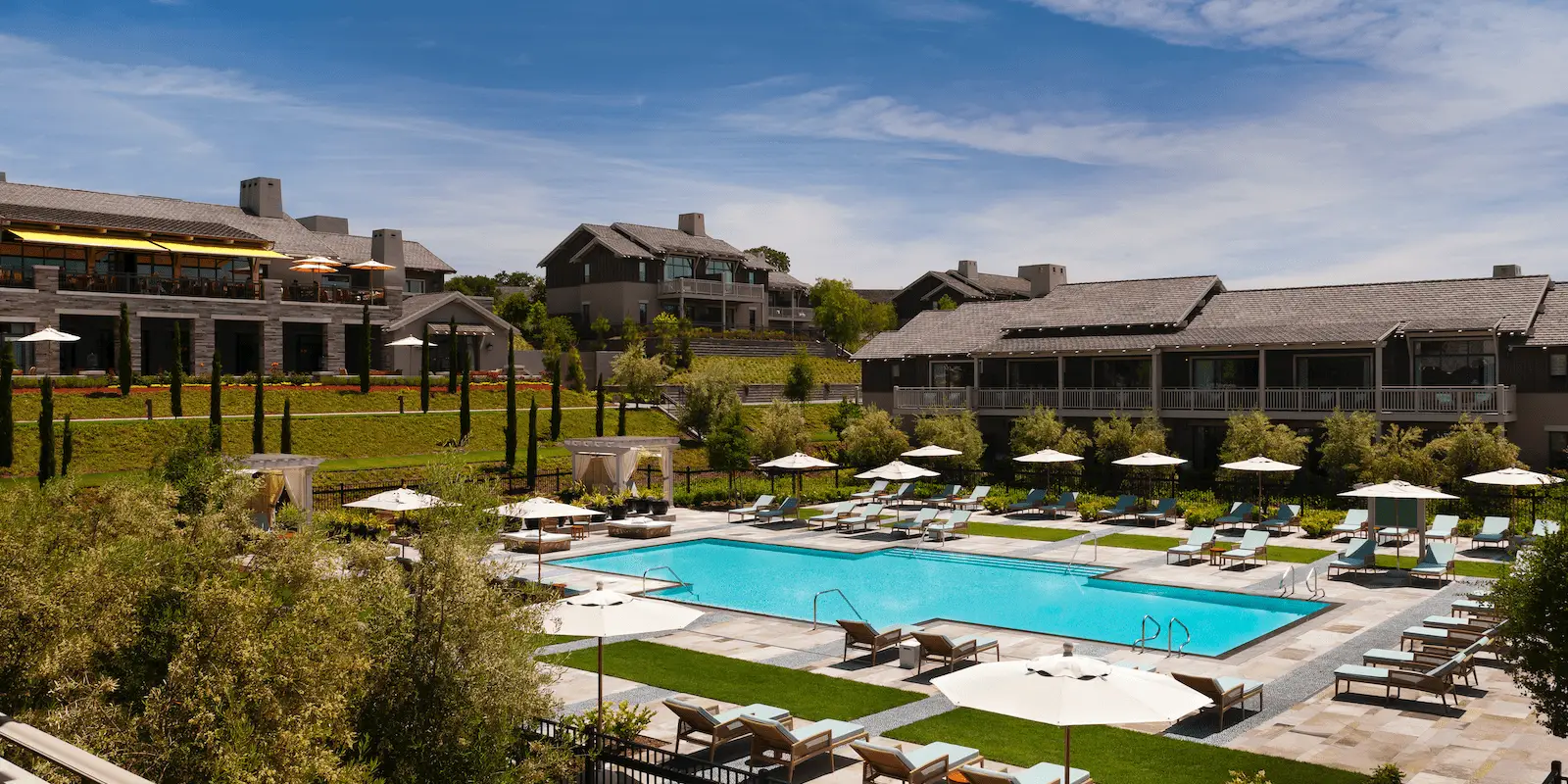 A luxurious resort, listed among the Best Spa and Wellness Hotels in the Bay Area, features a large outdoor swimming pool surrounded by lounge chairs, umbrellas, and lush greenery. Several buildings with balconies overlook the pool area. The sky is clear and blue, creating a serene and inviting atmosphere.