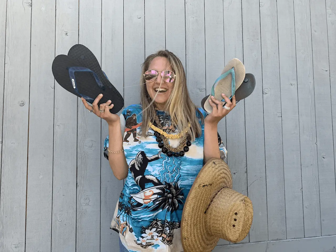 A woman stands against a wooden wall, smiling widely. She wears a colorful Hawaiian shirt, sunglasses, and a beaded necklace. Holding up a pair of flip-flops in each hand with a straw hat hanging on her left arm, she embodies the spirit captured by the top 13 Hawaiian Pidgin words to know.