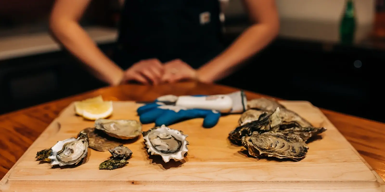 A person stands behind a wooden cutting board holding a shucking knife, wearing a blue glove. The board displays various opened oysters, an unshucked oyster, and a lemon wedge. The scene suggests preparation of fresh Sonoma Coast seafood.