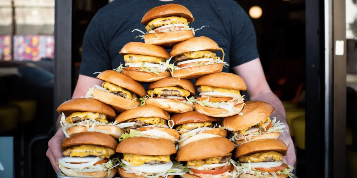 A person holds a towering stack of burgers. Each burger is layered with cheese, lettuce, tomatoes, onions, and sauces, all inside golden buns. The background is slightly blurred, indicating a restaurant setting known for the best burgers San Francisco has to offer.
