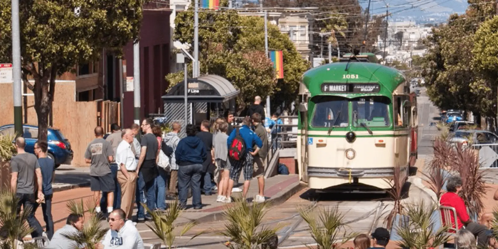 A group of people wait at a bus stop on a sunny day in San Francisco, set on a city street lined with trees and colorful buildings. A green and white streetcar approaches the stop, with passengers aboard and wires visible overhead. The area is bustling with activity, resembling some of the best parklets in San Francisco.