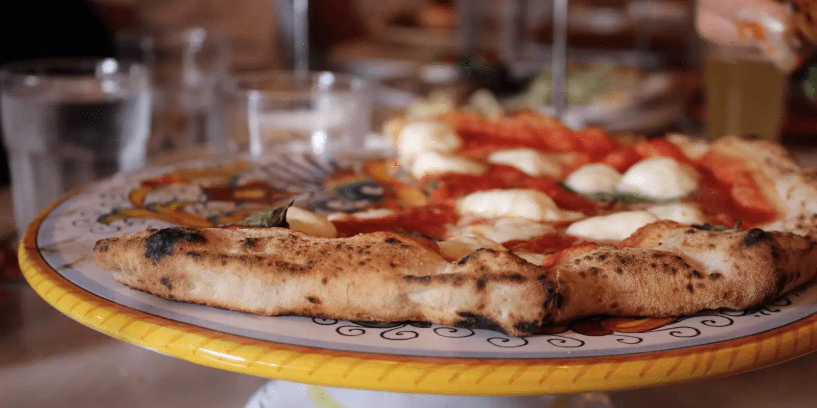 Close-up view of a margherita pizza on a decorative yellow plate. The pizza has a crispy crust, visible patches of melted mozzarella cheese, tomato sauce, and fresh basil. Blurry background shows glasses and plates, indicating a dining setting. Experience the best pizza San Francisco has to offer right at your table!