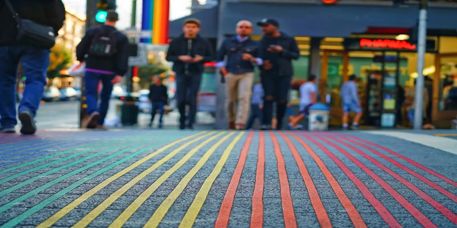 A group of people walk along a city street with a colorful rainbow-painted crosswalk in the foreground. The background shows buildings, shops, and other pedestrians, and a rainbow flag hanging above the street on the left, as they celebrate San Francisco Pride all month long with these special events.
