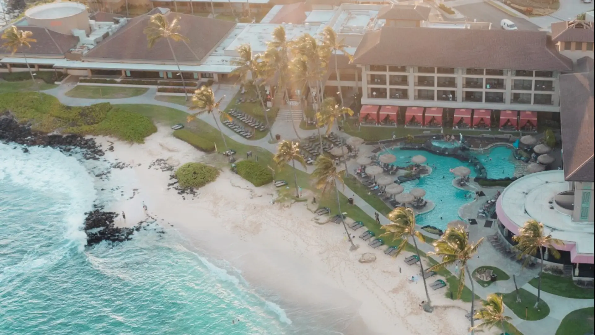 Aerial view of one of the best oceanfront hotels in Kauai, featuring a stunning beachfront resort with white sandy beach, turquoise ocean, and palm trees. The resort boasts multiple buildings, a large pool with lounge chairs and red umbrellas. Waves gently crash on the shoreline, surrounded by lush green landscaped areas.