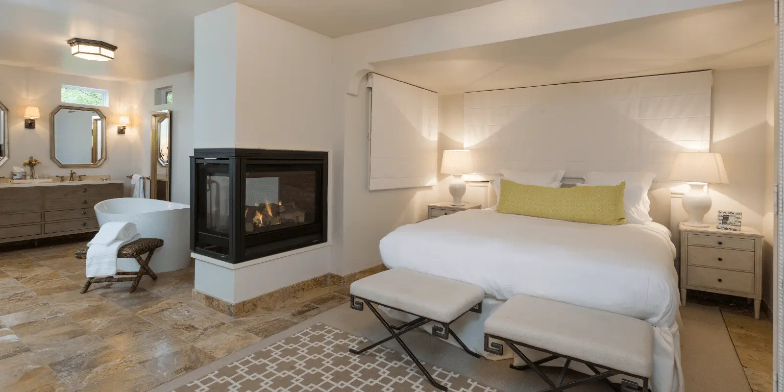 A spacious bedroom with a double bed featuring a green bolster pillow, flanked by nightstands and lamps. There's a cozy fireplace next to the bed, and an adjoining bathroom with a freestanding bathtub, double sinks, mirrors, and pendant lighting. Reminiscent of the best spa wellness hotels in Sonoma, neutral tones throughout.