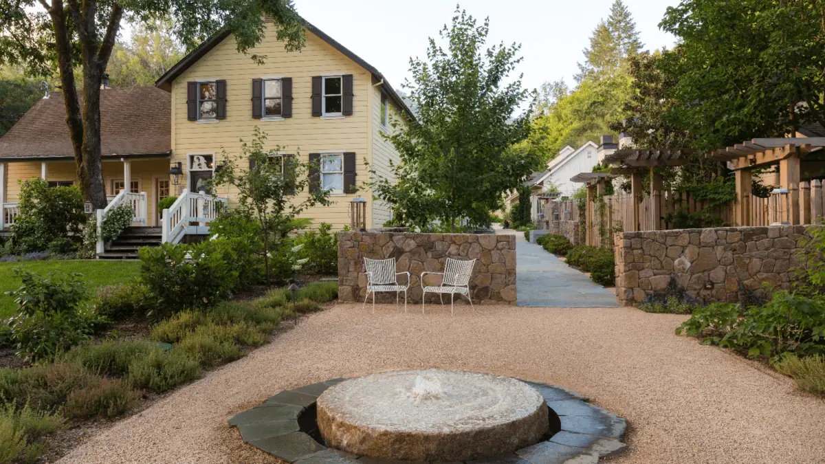 A stone-paved garden features a central water fountain and two white metal chairs. It is bordered by lush greenery and quaint houses, including a prominent yellow house with black shutters on the left. Reminiscent of architecturally iconic California hotels, a stone wall separates the garden from a tree-lined pathway.