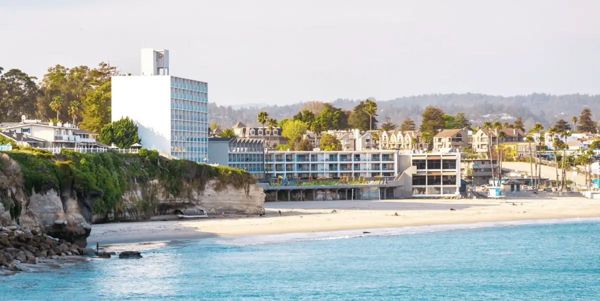 A scenic view of a beach with turquoise waters and a sandy shoreline. There are cliffs to the left with greenery on top. Four Santa Cruz hotels to put on your radar line the beachfront, with trees and hills visible in the background under a clear sky.