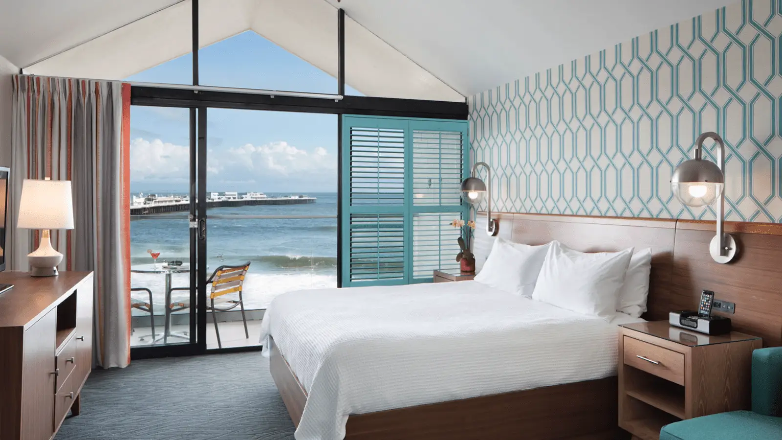 A modern hotel room with a king-sized bed, wooden furniture, and a geometric patterned accent wall. The room leads out to a balcony with a table and chairs, overlooking a scenic ocean view with waves and a pier in the distance—one of the Four Santa Cruz Hotels to put on your Radar.