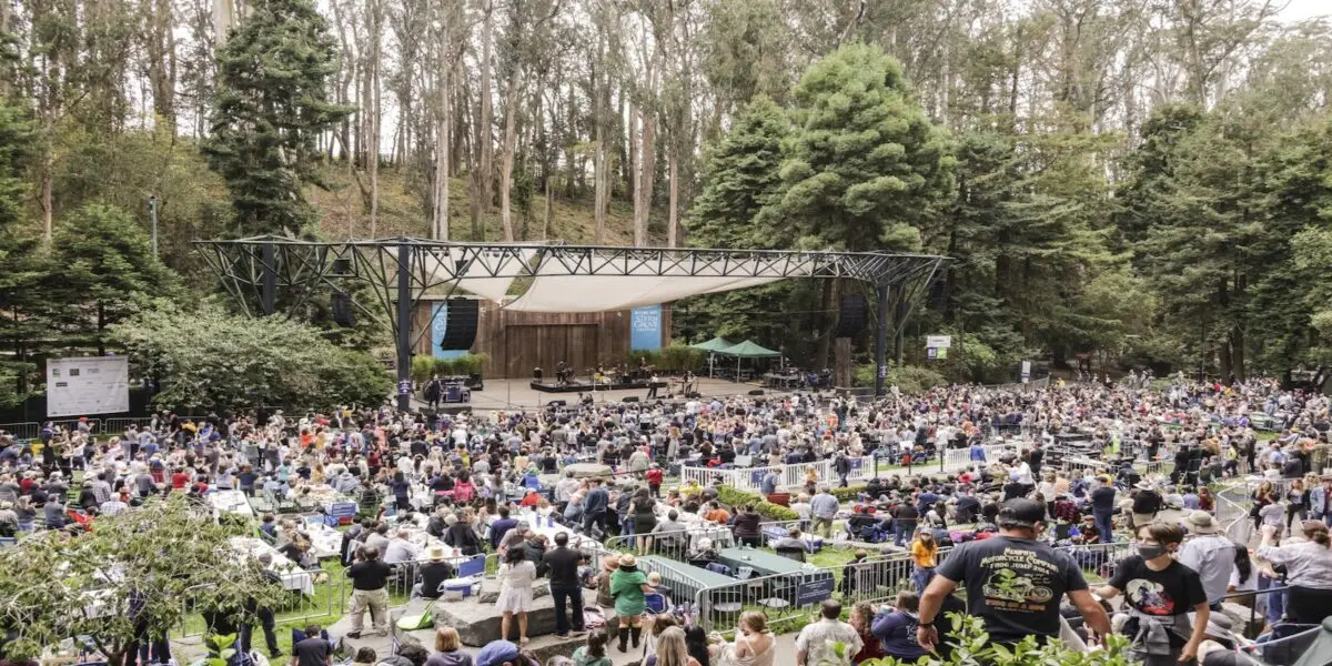 A large crowd gathers outdoors at an amphitheater surrounded by tall trees and a lush forest. Attendees are seated in front of a stage, which features a simple backdrop and a canopy overhead. The atmosphere is lively, with people chatting and moving around—a perfect addition to your things to do Bay Area June list.