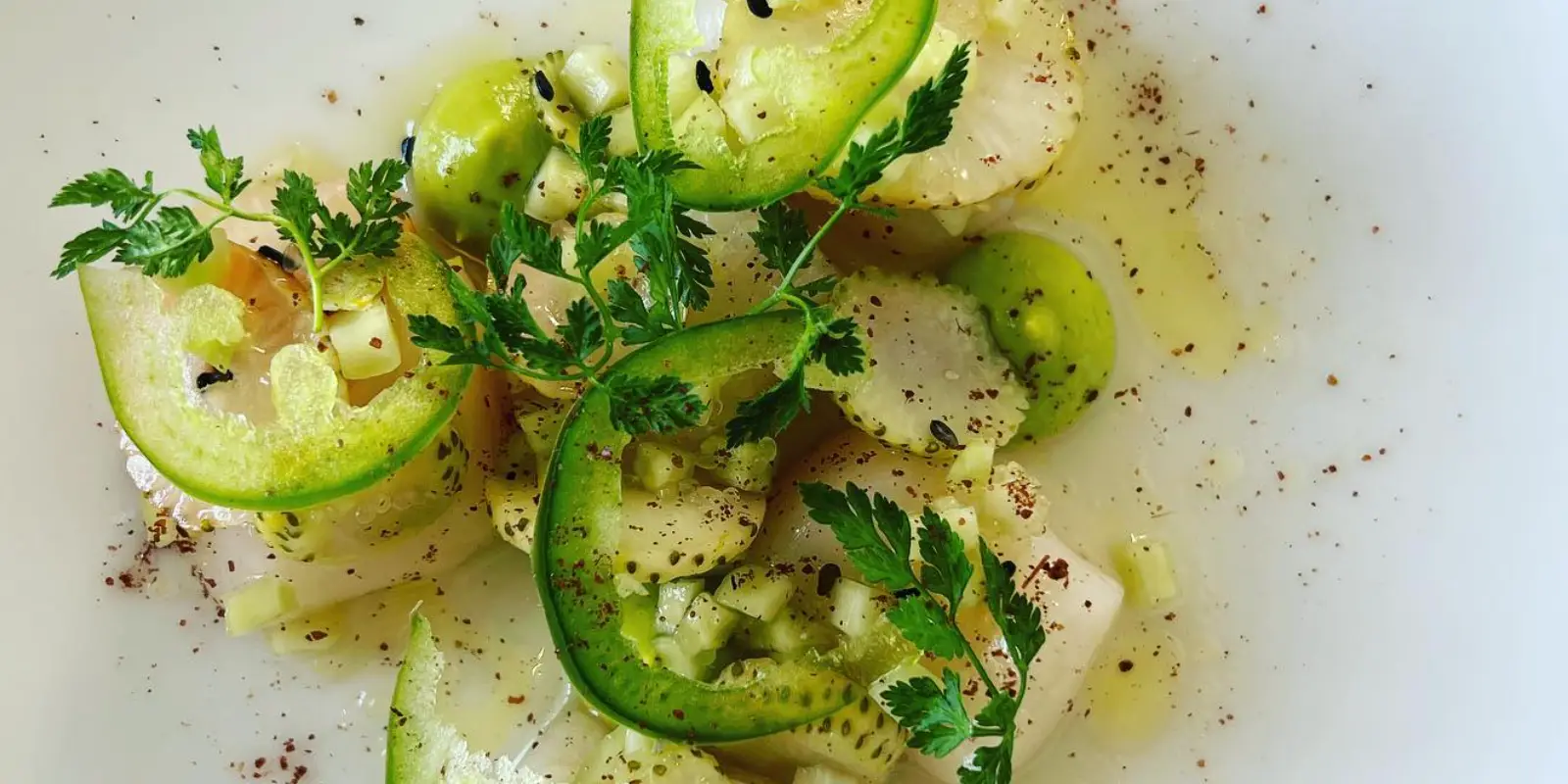 A close-up image of a fresh salad dish featuring thinly sliced cucumbers, green tomatoes, and garnished with herbs. Seasoned with black pepper and a drizzle of olive oil, it has a vibrant and appetizing appearance on a white plate—certainly one of the best breakfast options on the Monterey Peninsula.