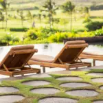 Three wooden lounge chairs with brown cushions face a tranquil infinity pool. The pool overlooks a lush, green golf course and distant palm trees. Large circular stepping stones embedded in grass lead up to the pool area at one of the best luxury hotels Kauai offers, creating a serene and inviting scene.