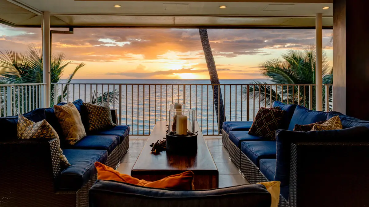 A cozy outdoor seating area with blue cushioned chairs and a central wooden table, arranged on a balcony overlooking the ocean at sunset. Palm trees frame the view, and decorative candles sit on the table, creating a serene and inviting atmosphere—perfect for those staying at the best group hotels in Maui.