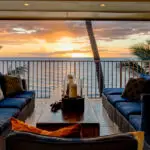 A cozy outdoor seating area with blue cushioned chairs and a central wooden table, arranged on a balcony overlooking the ocean at sunset. Palm trees frame the view, and decorative candles sit on the table, creating a serene and inviting atmosphere—perfect for those staying at the best group hotels in Maui.