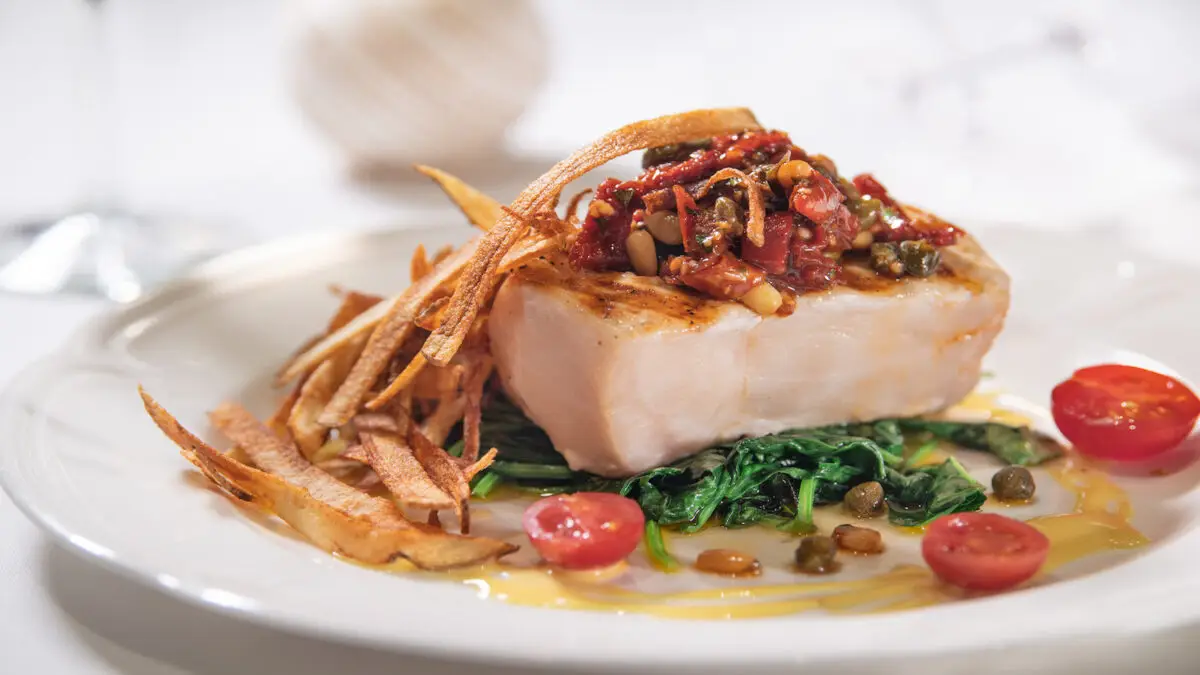 A gourmet dish featuring a seared piece of fish topped with a tomato and pine nut relish, served on a bed of sautéed greens. Crispy fried strips are placed on the side, and cherry tomatoes with capers garnish the plate. Elegantly plated on white china, it's among the best dinners on the Monterey Peninsula.