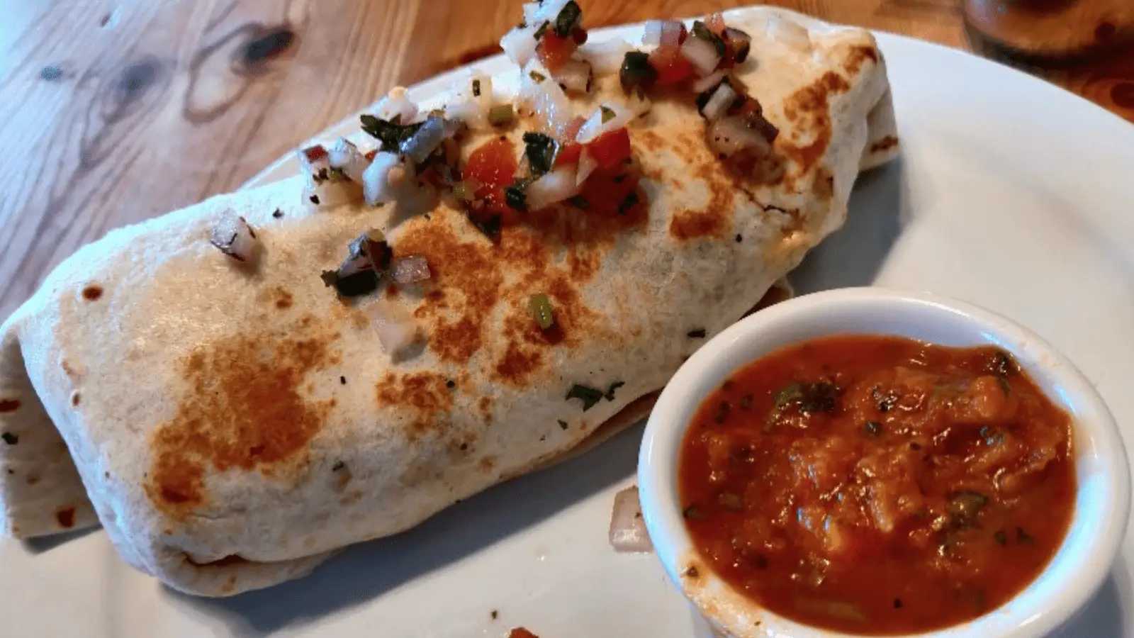 A burrito topped with salsa and herbs is placed on a white plate. Next to the burrito is a small white bowl filled with chunky red salsa. The plate, perfect for the best breakfast on the Monterey Peninsula, is set on a wooden table.