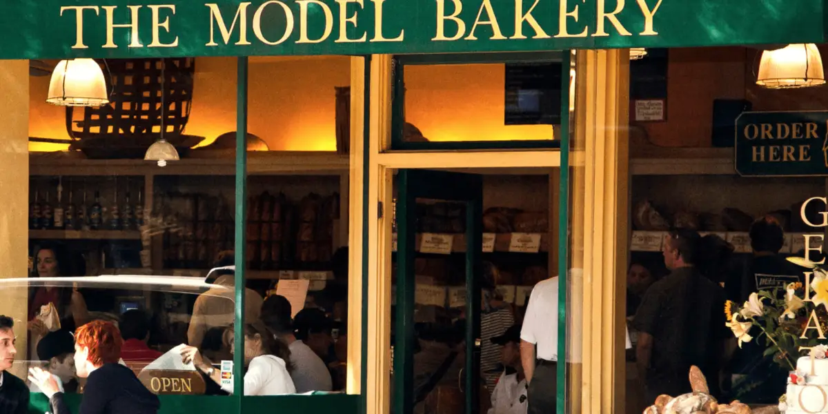 A busy bakery storefront with a green and yellow sign reading "The Model Bakery." Patrons are seen inside through large windows, some seated and others standing in line. The interior is warmly lit, and loaves of bread are displayed on shelves in the background, offering the best breakfast in Wine Country.