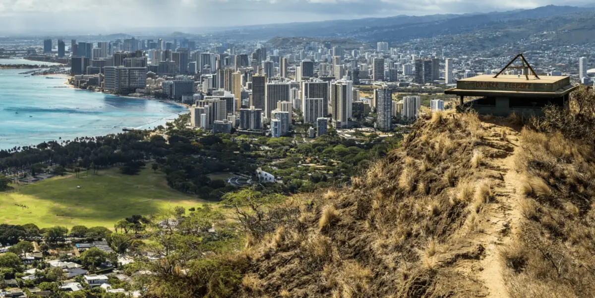 View from Diamond Head Summit looking down on Waikiki, Honolulu. The image shows a hiking trail leading to a bunker with views of the city skyline, beach, and ocean. This spot is often highlighted in the best island tours of Oahu for its lush green spaces contrasting with the urban landscape.