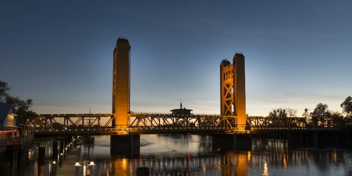 A nighttime view of the Tower Bridge, one of the iconic California bridges, illuminated with golden lights in Sacramento. The bridge features two prominent towers connected by a central span reflecting on the calm river below. The sky is a dusky blue with a hint of dusk's last light.