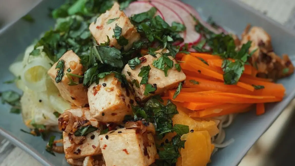 A colorful salad is served with cubes of seasoned tofu, shredded carrots, cilantro, radishes, and greens on a bed of noodles. Reminiscent of the best Hawaiian food in Kauai, the marinated tofu with herbs and spices adds a flavorful touch to the fresh vegetables.