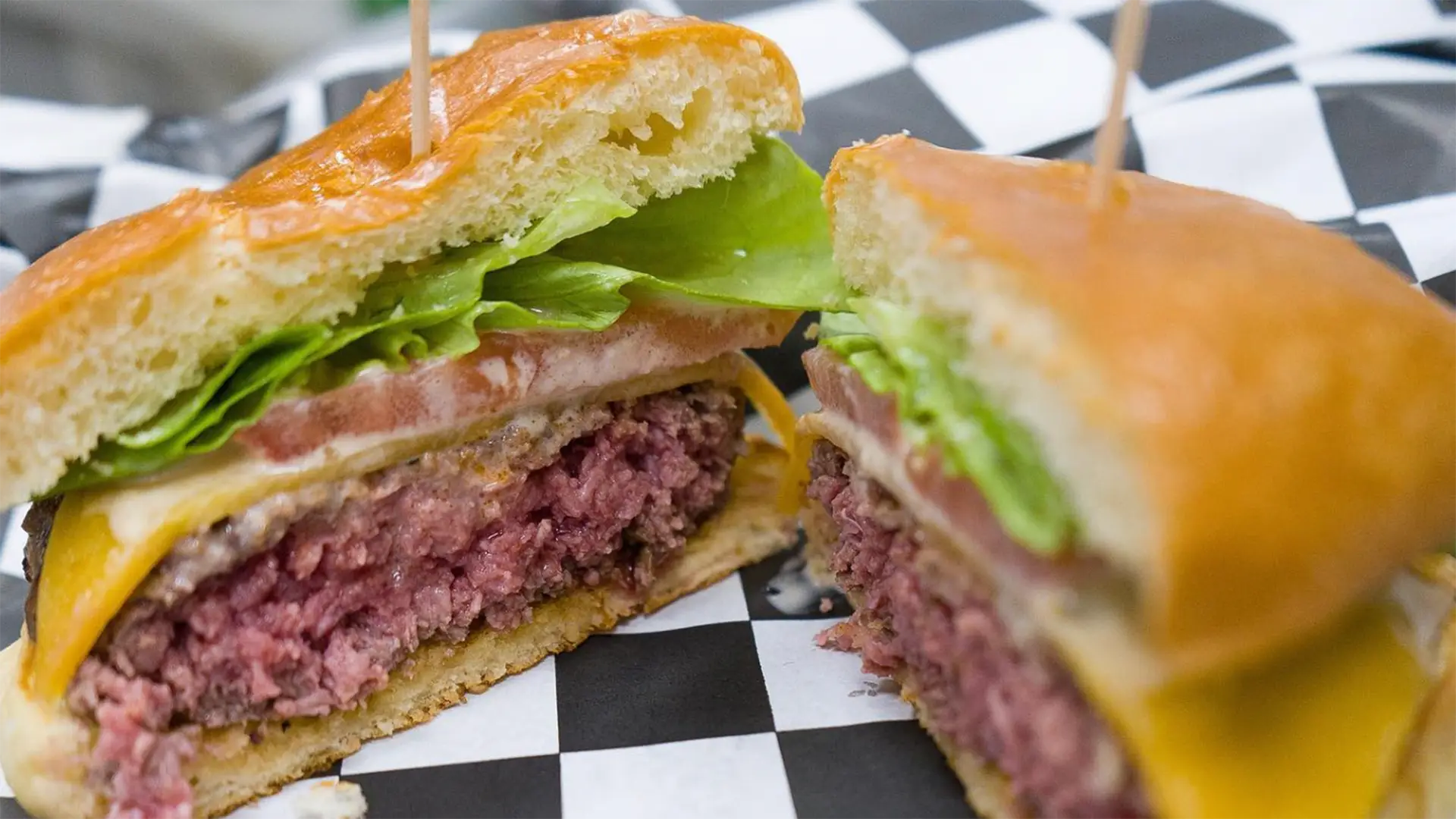 A close-up of a hamburger cut in half, revealing a juicy pink patty, melted cheese, tomato slices, and lettuce inside a shiny, golden bun. The halves are placed on a black and white checkered paper, held together with toothpicks. This could easily be the best burger on the Big Island.