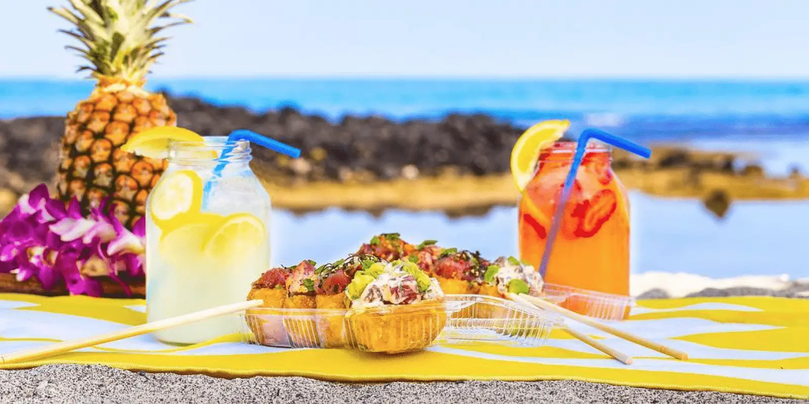 A beach picnic setup for the best happy hour on the Big Island, featuring two colorful mason jar drinks—one yellow and one red—with blue straws and lemon slices. In the center, a tray of appetizers garnished with herbs. There's a pineapple and purple flowers in the background, with the ocean visible.