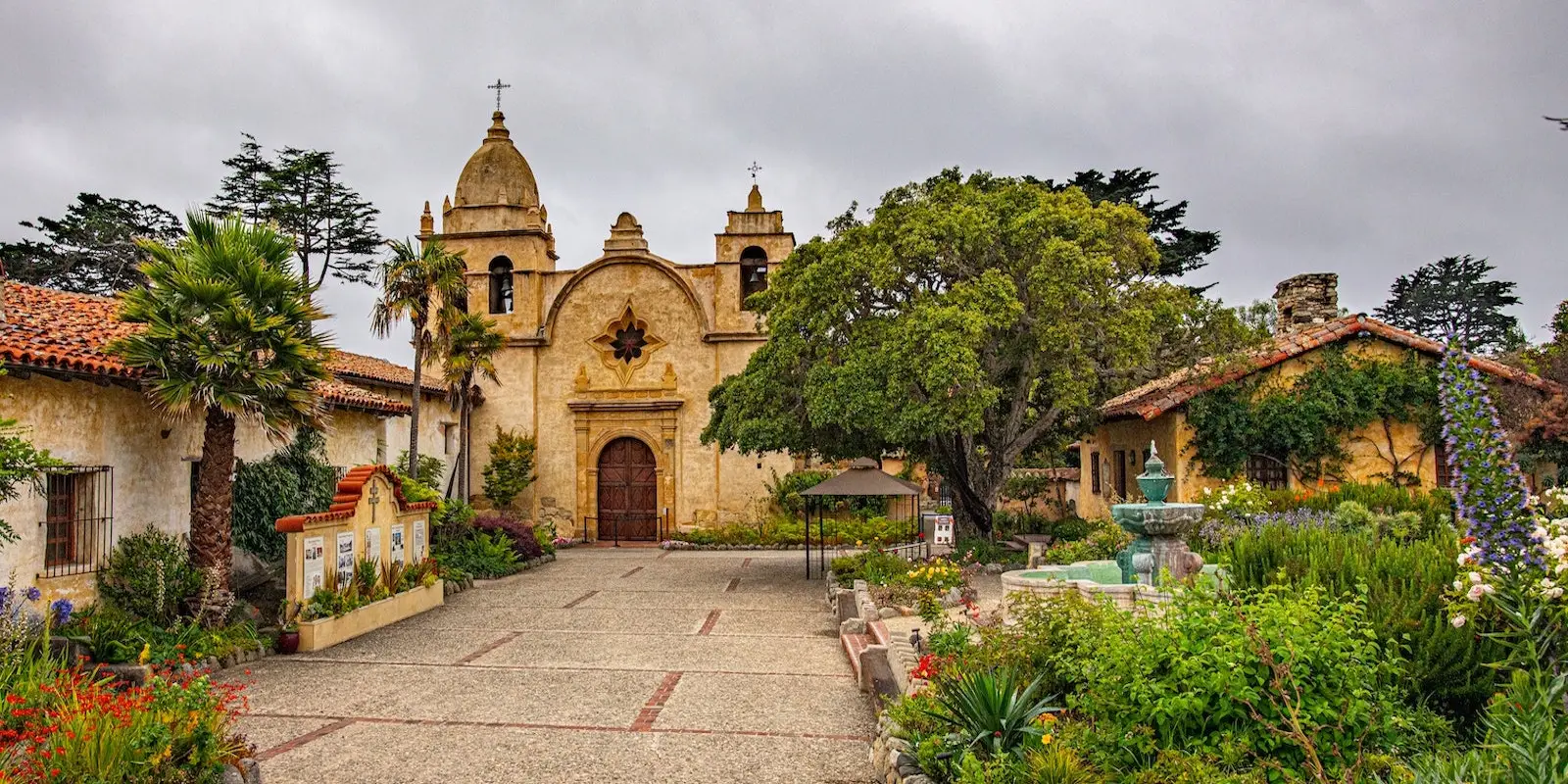 The image depicts Mission San Carlos Borromeo de Carmelo in California. The historic Spanish colonial church, often highlighted in the top self-guided tours in Carmel, features a stone facade with two bell towers, a central wooden door, and surrounding lush gardens with a fountain. Overcast sky in the background.