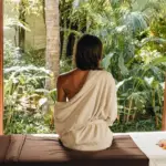 A person with long hair sits on a massage table wrapped in a towel at the best spa hotel in Maui, facing a lush garden with various tropical plants and trees. A window on both sides frames the relaxing scene, and there are flowers placed on the table beside the person.