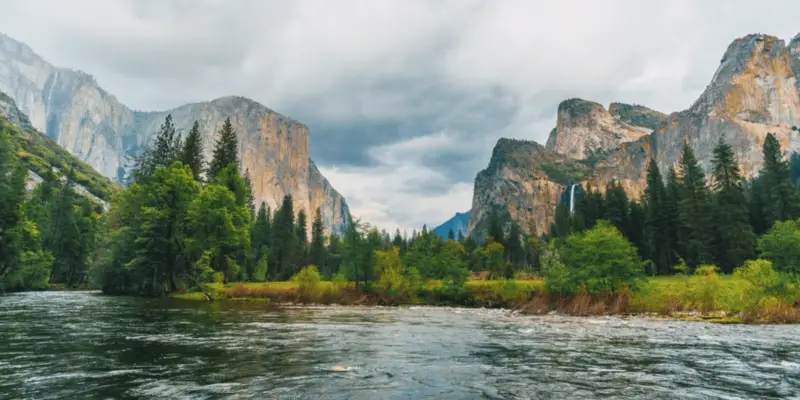 A flowing river runs through a lush valley surrounded by steep, rocky mountains and dense forests under a cloudy sky. The greenery is vibrant, and the landscape is majestic, depicting a serene and picturesque natural scene reminiscent of Yosemite National Park.