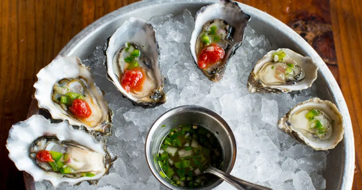 A plate of six raw oysters on the half shell, arranged on a bed of crushed ice. Each oyster is garnished with chopped green onions, red roe, and a small tin cup of green mignonette sauce in the center. Perfectly presented for the best lunch San Francisco has to offer, all set on a wooden table.