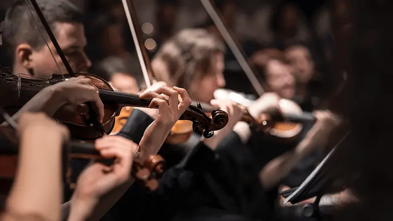 Close-up of several musicians playing violins in an orchestra. The hands and instruments are in focus, while the faces of the musicians are blurred, highlighting the concentration and intricacy of the performance. Experiencing such a mesmerizing concert is one of the best things to do on Monterey Peninsula in July.