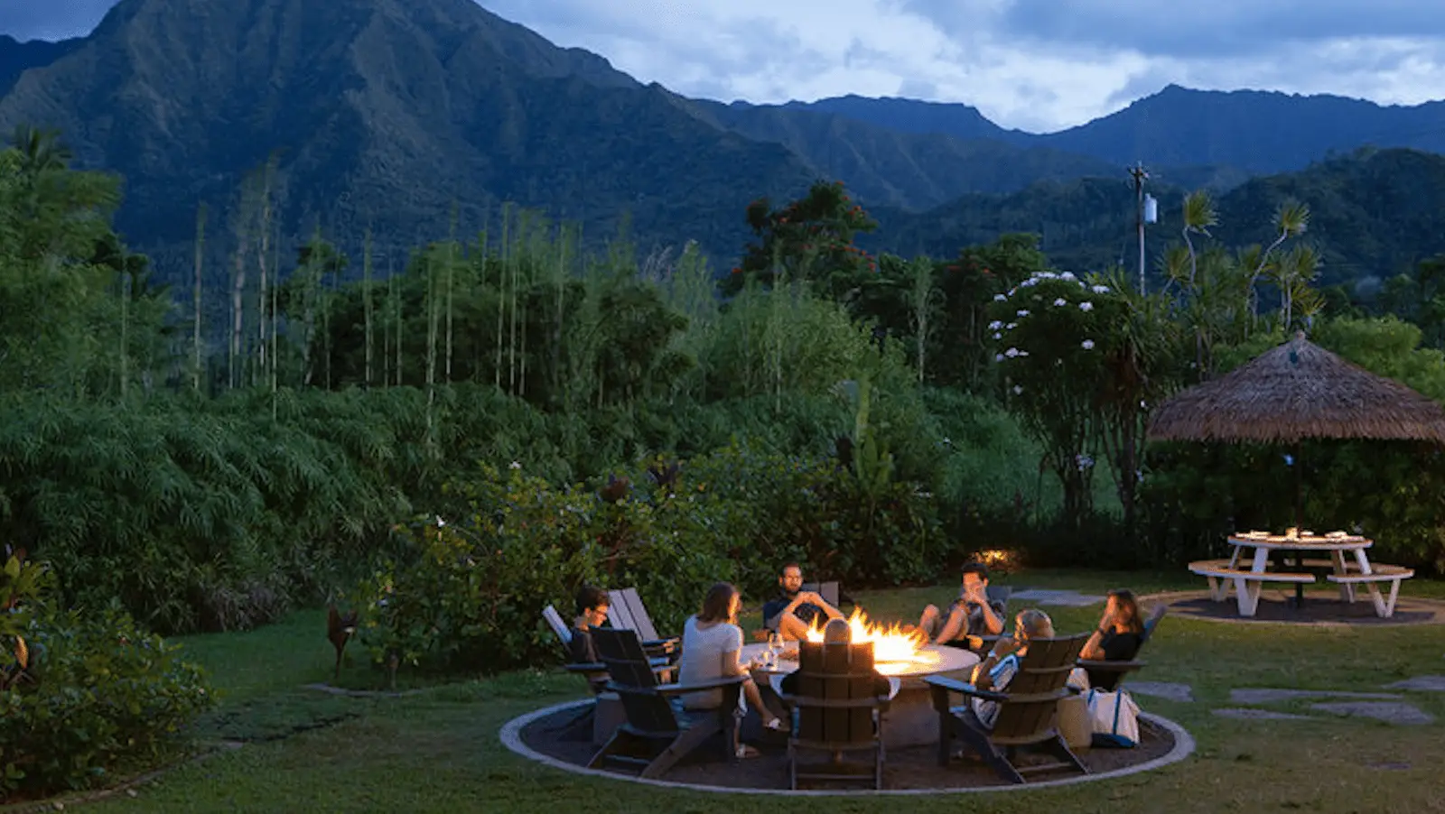 A group of people sit around a fire pit at dusk, the sky lit with the hues of sunset, surrounded by lush greenery and mountains in the background. It’s an idyllic setting for their Kauai retreat. Some engage in conversation while others relax in Adirondack chairs, enjoying the serene ambiance of their dinner gathering.