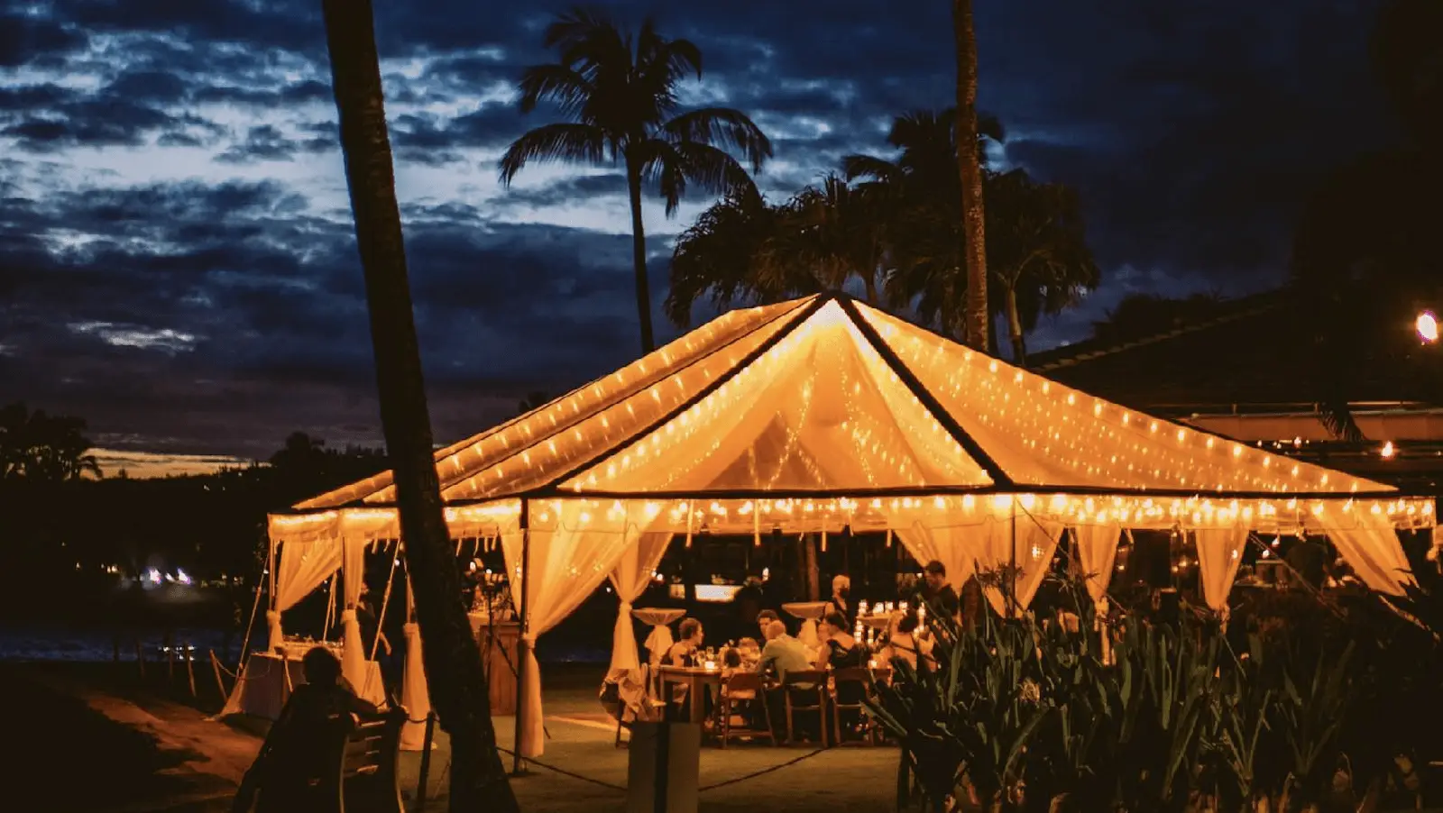 An outdoor sunset dinner event at night under a warmly-lit pavilion draped with white fabric and string lights. People are seated at tables inside, surrounded by palm trees and lit by soft, ambient lighting. The sky over Kauai is dark with hints of an evening glow.