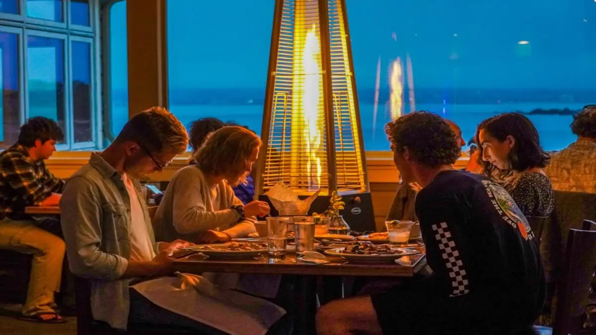 A group of people sitting around a table at a cozy restaurant, considered to have the best restaurant view on the Monterey Peninsula, are illuminated by an outdoor heater. They are enjoying dinner while looking at their food and conversing. The background shows large windows with a view of the evening sky and a body of water.