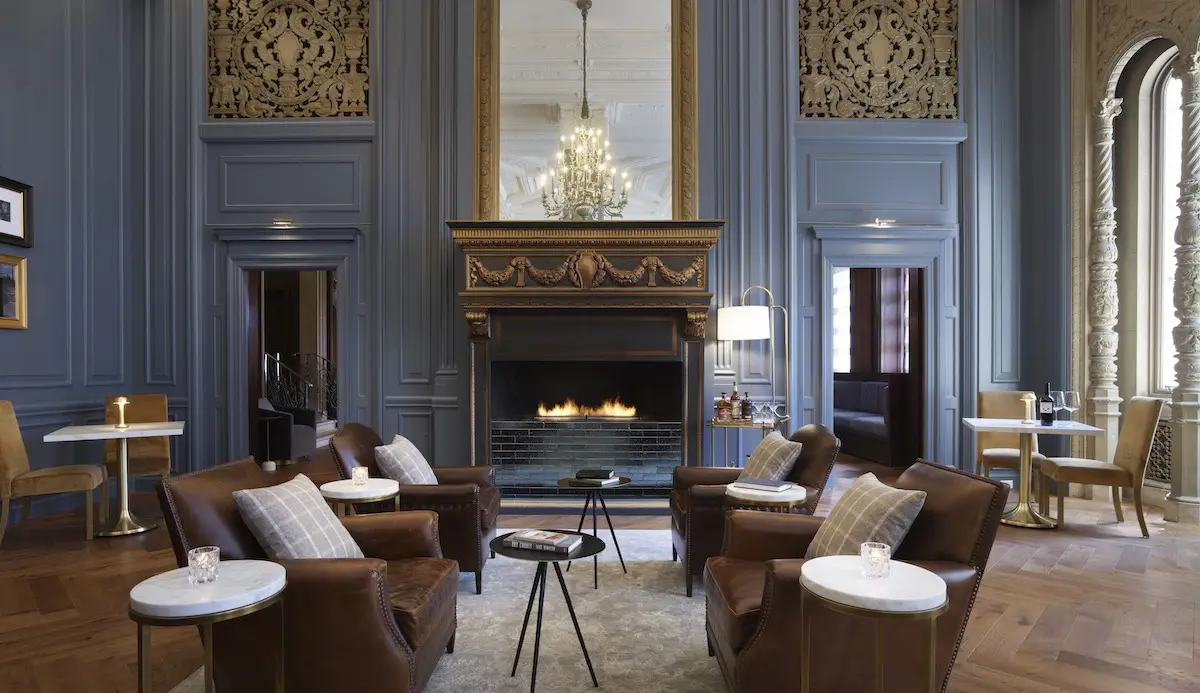 A luxurious lounge at the Union Square Hotel features four leather armchairs with plaid pillows arranged around a small circular table and a rug, in front of an ornate fireplace. The room boasts high ceilings, intricate wall details, large mirrors, a chandelier, and multiple seating areas.