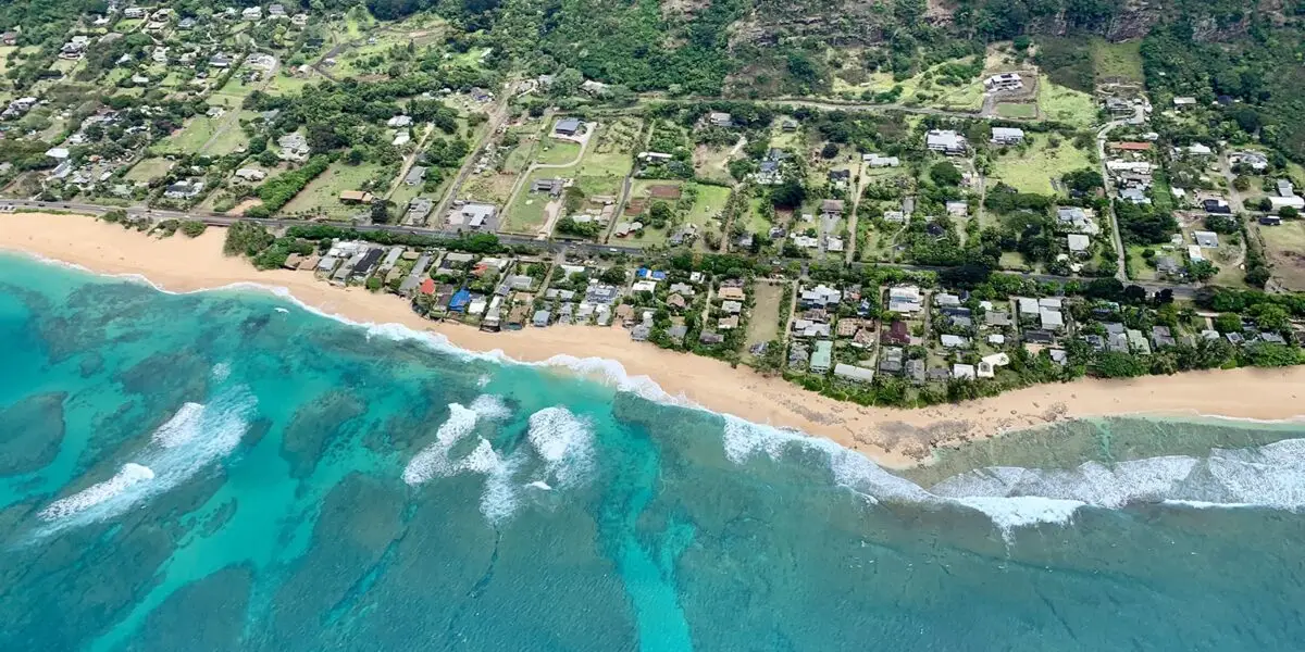 Aerial view of a coastal town featuring a sandy beach with turquoise waters and white waves. The town has numerous houses surrounded by greenery and vegetation, situated at the edge of a forested area with hills in the background. It feels like experiencing the best helicopter tour Oahu has to offer.