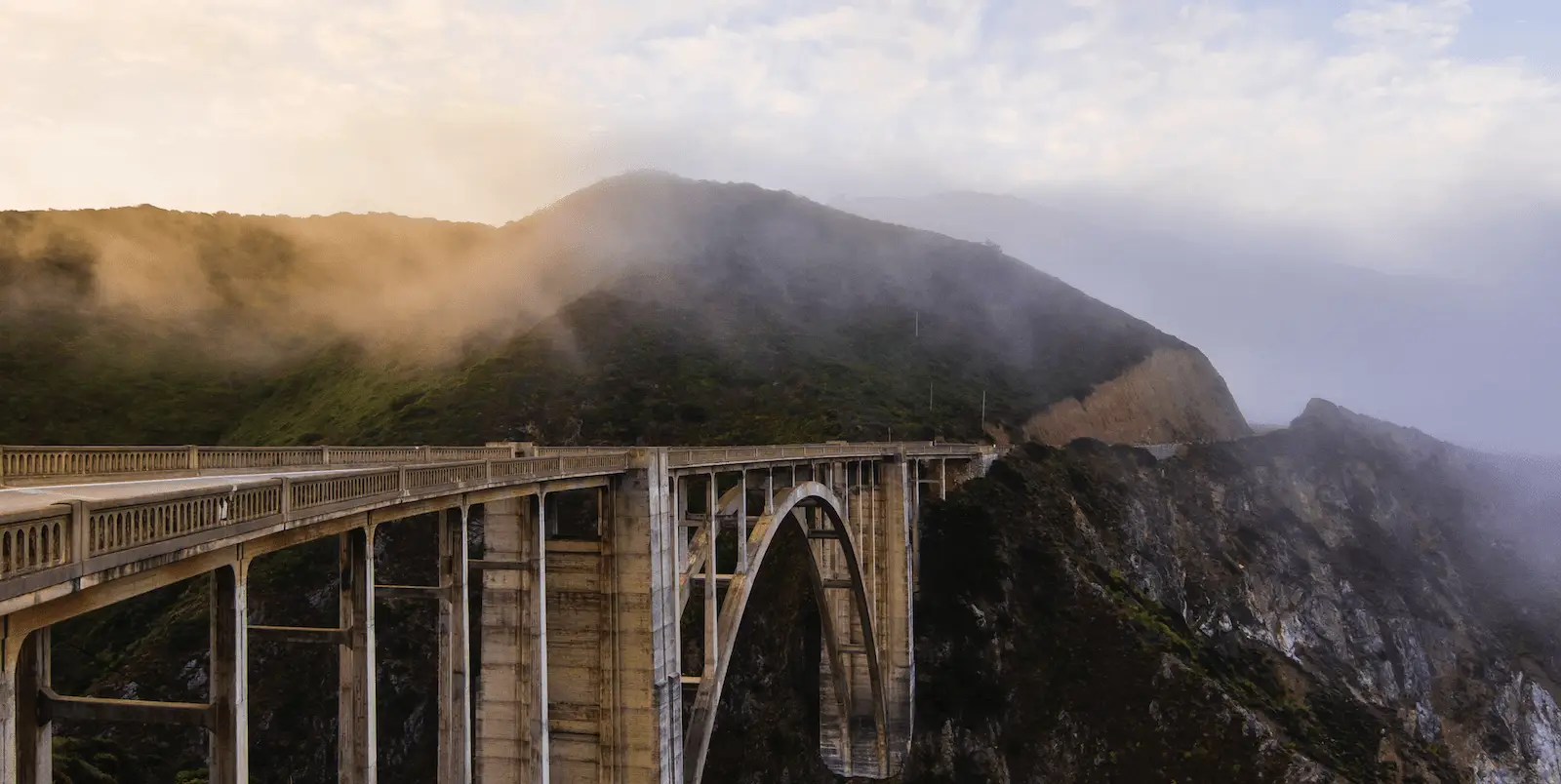 A picturesque view of a concrete arch bridge spanning a craggy cliffside, with fog rolling over the lush green hills in the background, under a sky filled with soft twilight hues. This iconic sight is among the highlights of what to do on the Monterey Peninsula, showcasing nature's beauty and engineering marvels.