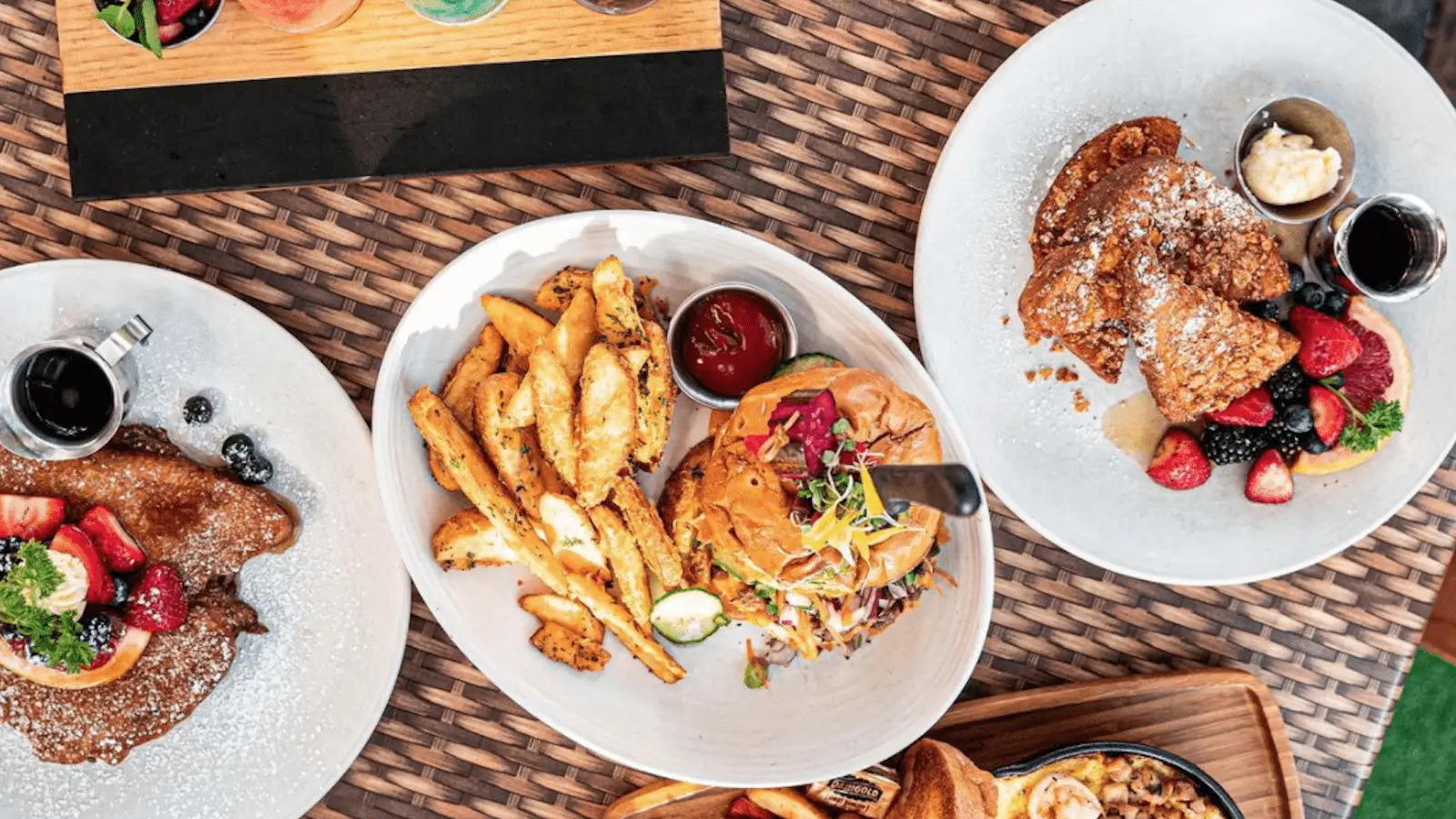 Top-down view of a table with assorted dishes. Includes a plate of crispy fries and a burger, French toast topped with powdered sugar and berries, and pancakes with fruit. Small containers of sauces and syrups are also on the table. The surface is wicker-style.