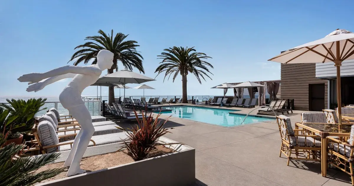 Outdoor poolside area with lounge chairs, umbrellas, and tables shaded by palm trees. A white statue of a human figure in a diving pose is positioned near the edge of a seating area. Clear blue sky and ocean visible in the background—truly one of the best hotel pools in California.