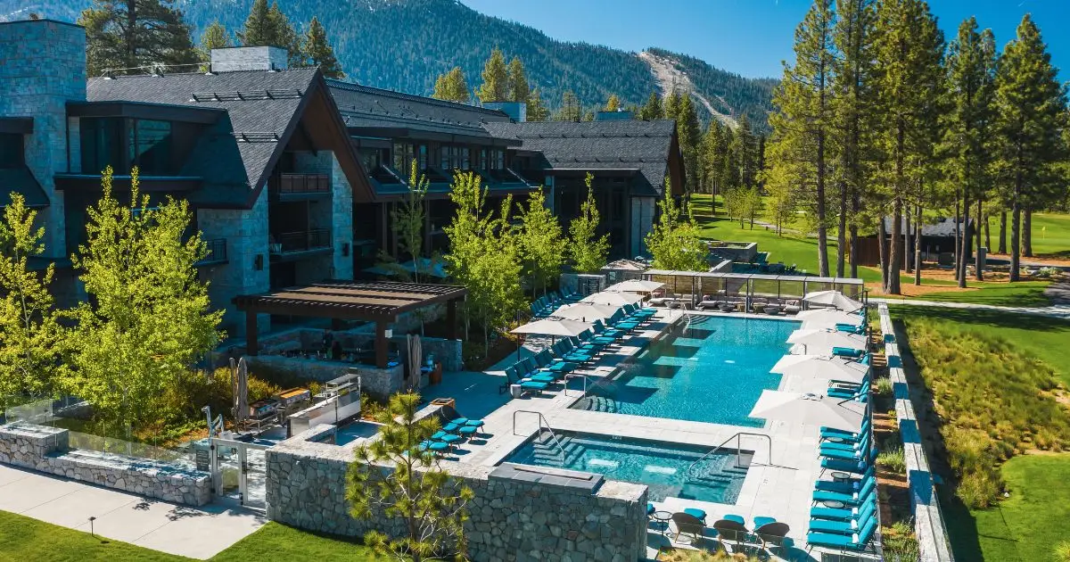 A luxurious mountain resort, one of the best hotel pools in California, features a central outdoor swimming pool surrounded by lounge chairs and umbrellas. The stone structures and modern architecture blend with lush greenery and tall trees, offering a serene view of distant forested mountains.