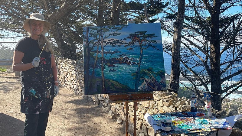 Painter stands next to coastal scene she painted for Carmel Art Walk on Monterey Peninsula, California.