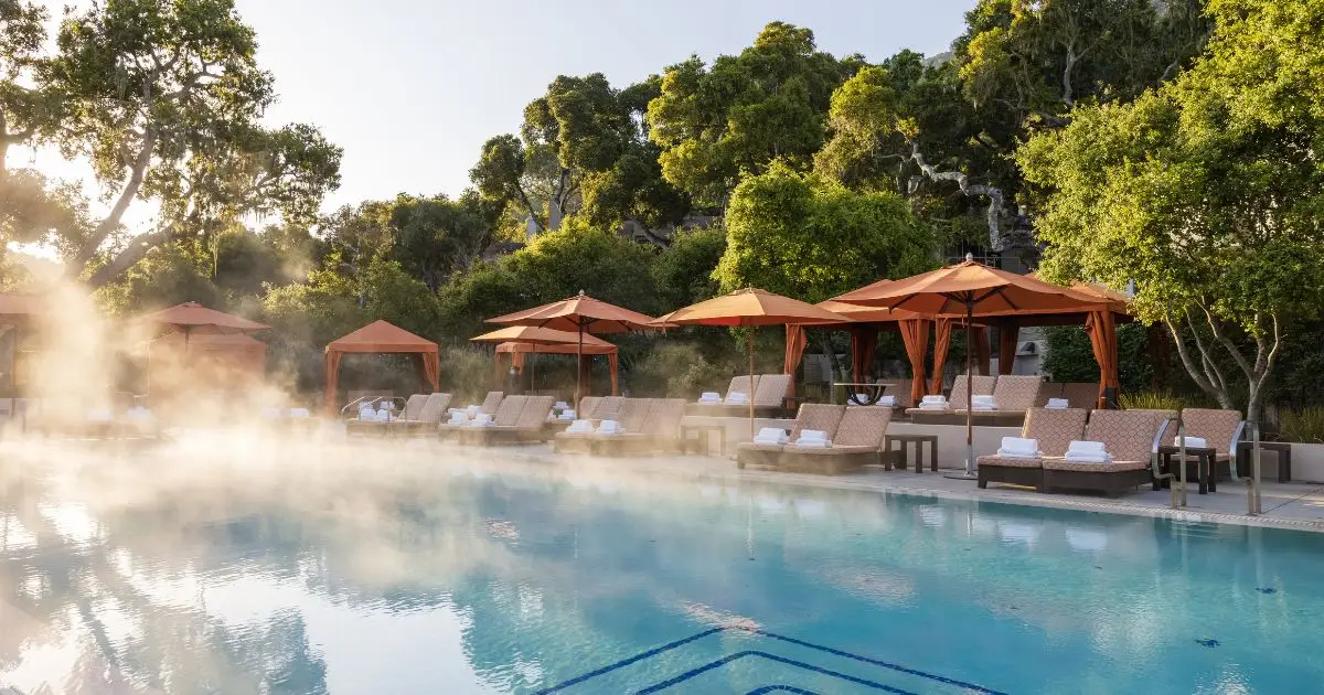 A serene outdoor pool area, often cited among the best hotel pools in California, features cushioned lounge chairs and stacked towels shaded by orange umbrellas. Behind, lush green trees provide a natural backdrop. Steam rises gently from the pool, suggesting warm water in a tranquil setting.