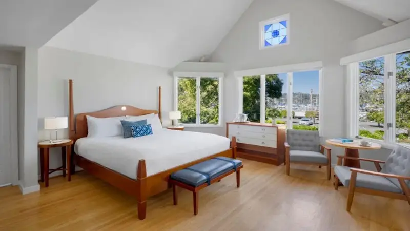 A bright bedroom in one of the finest luxury hotels in the Bay Area features a large bed covered in white linens and a blue accent pillow. It includes wooden furniture such as bedside tables, a chest of drawers, and a bench. Large windows provide views of lush trees and a marina. Near the window, two chairs and a small table await your relaxation.
