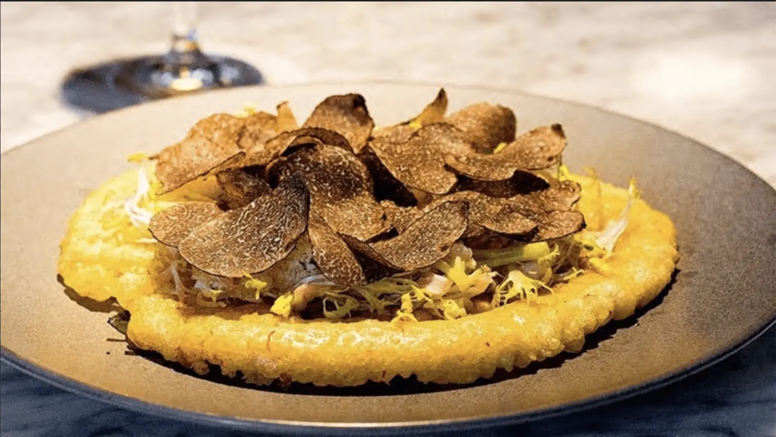 A culinary dish featuring a crispy potato pancake topped with generous slices of black truffle, served on a dark plate. The background is blurred, with a wine glass partially visible.