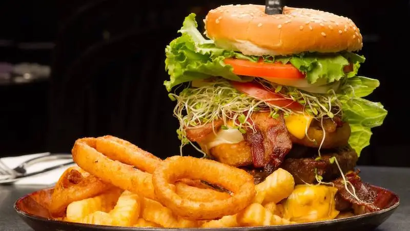 Close-up of a loaded cheeseburger topped with lettuce, tomato, sprouts, bacon, and a sesame seed bun on a plate. The plate also contains crinkle-cut fries and several golden-brown onion rings. A knife is stuck through the burger to hold it together—perfect for the best happy hour on the Big Island.