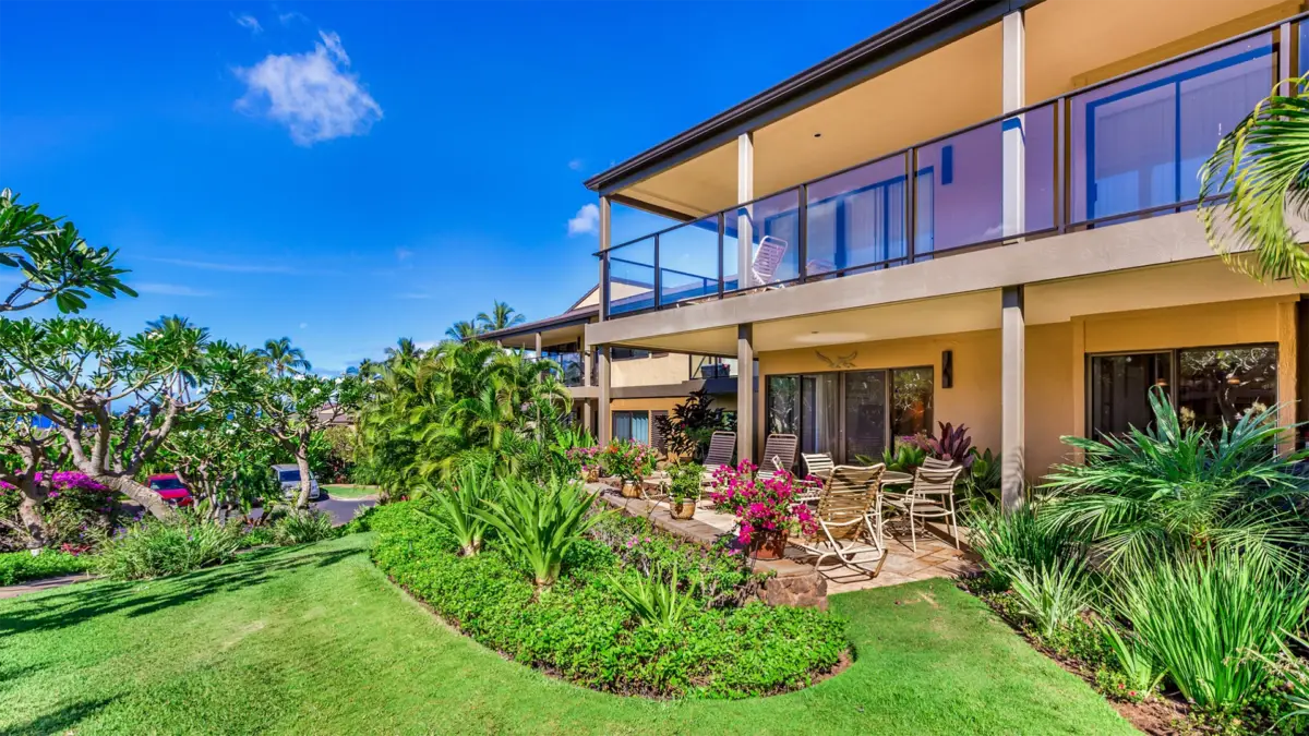 A two-story house with a balcony and patio, surrounded by lush greenery and tropical plants, reminiscent of the best hotels on Maui. Outdoor furniture is arranged on the patio beneath the balcony. The sky is clear and blue, enhancing the vibrant, sunny ambiance.